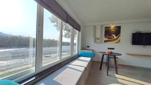 Een moderne kamer met een groot raam met uitzicht op een rivier, een bank met blauwe kussens, een kleine tafel met een schaal, en een aan de muur bevestigde televisie.