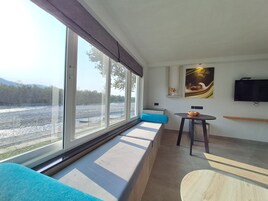 Una habitación moderna con un ventanal grande que da a un río, un sofá con cojines azules, una mesita con un bol y un televisor montado.