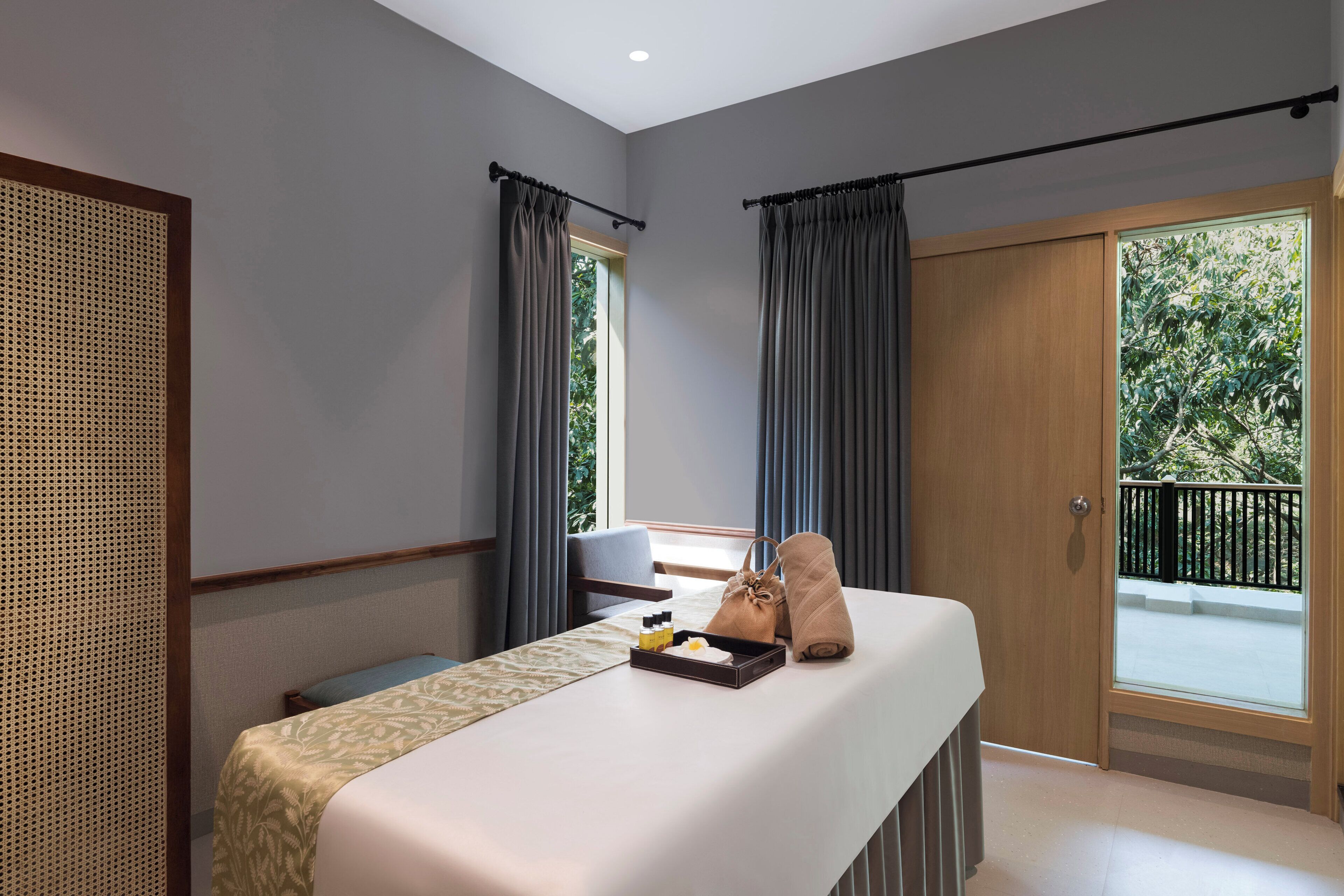 A hotel room with a bed, a small table, a window with curtains, and a balcony with a railing.