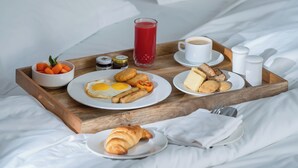 Een net opgemaakt bed met witte lakens, een houten dienblad met ontbijtartikelen, waaronder eieren, toast en een drankje, en een blauw hoofdeinde.