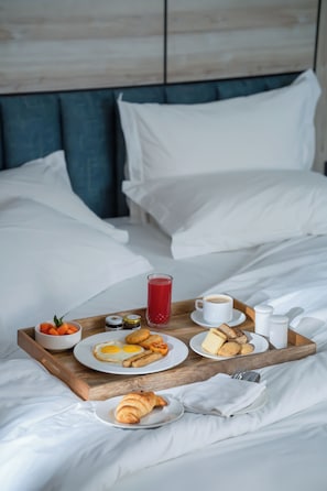 Cama bem arrumada com roupa de cama branca, bandeja de madeira com itens de café da manhã incluindo ovos, torradas e uma bebida, e cabeceira azul.