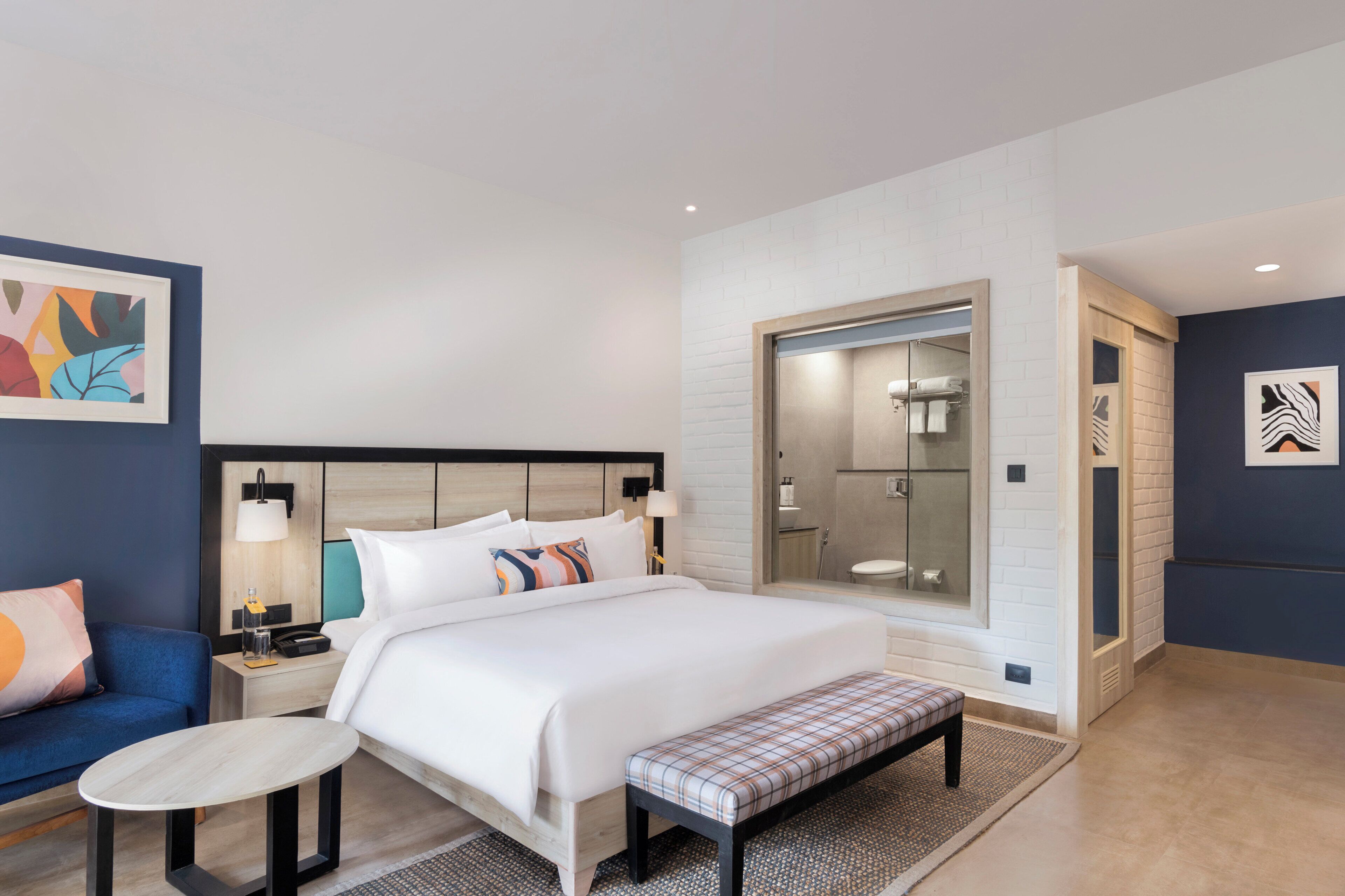 A modern hotel room with a large bed, bedside tables, a bench, a small table, a shower area, and artwork on the walls.