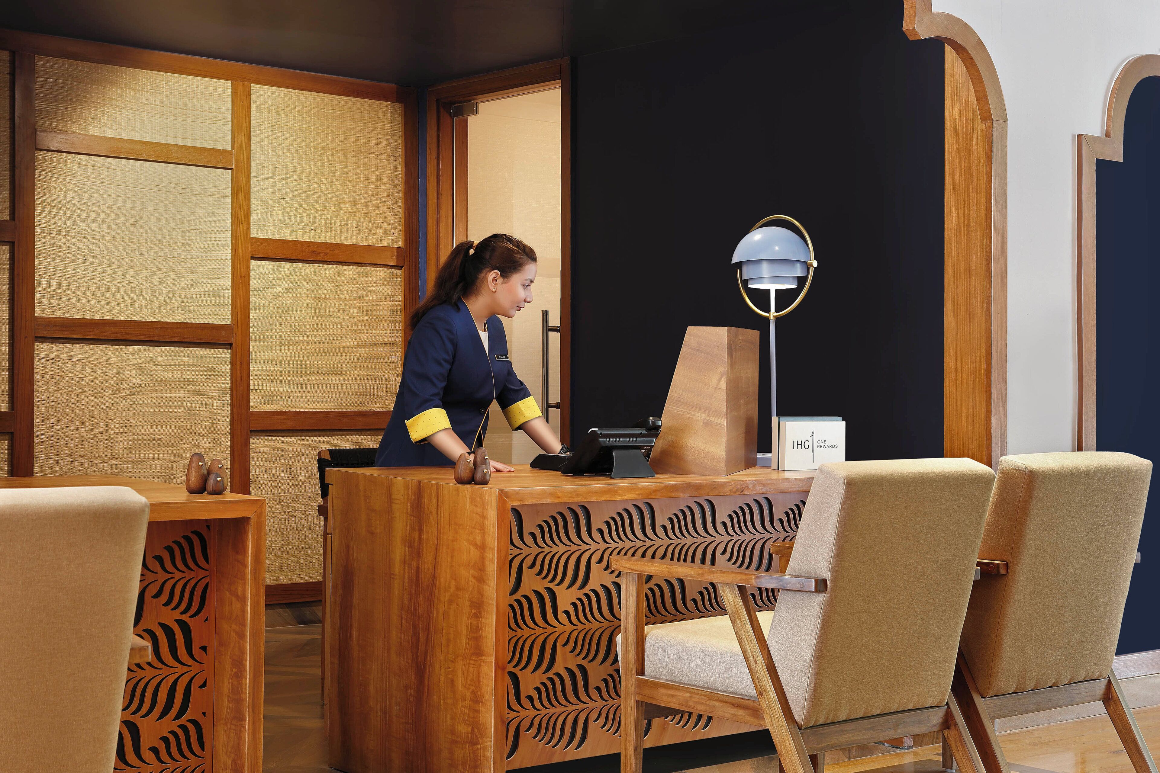Un comptoir d’accueil au design en bois, une lampe moderne et un espace détente avec des fauteuils à motifs.
