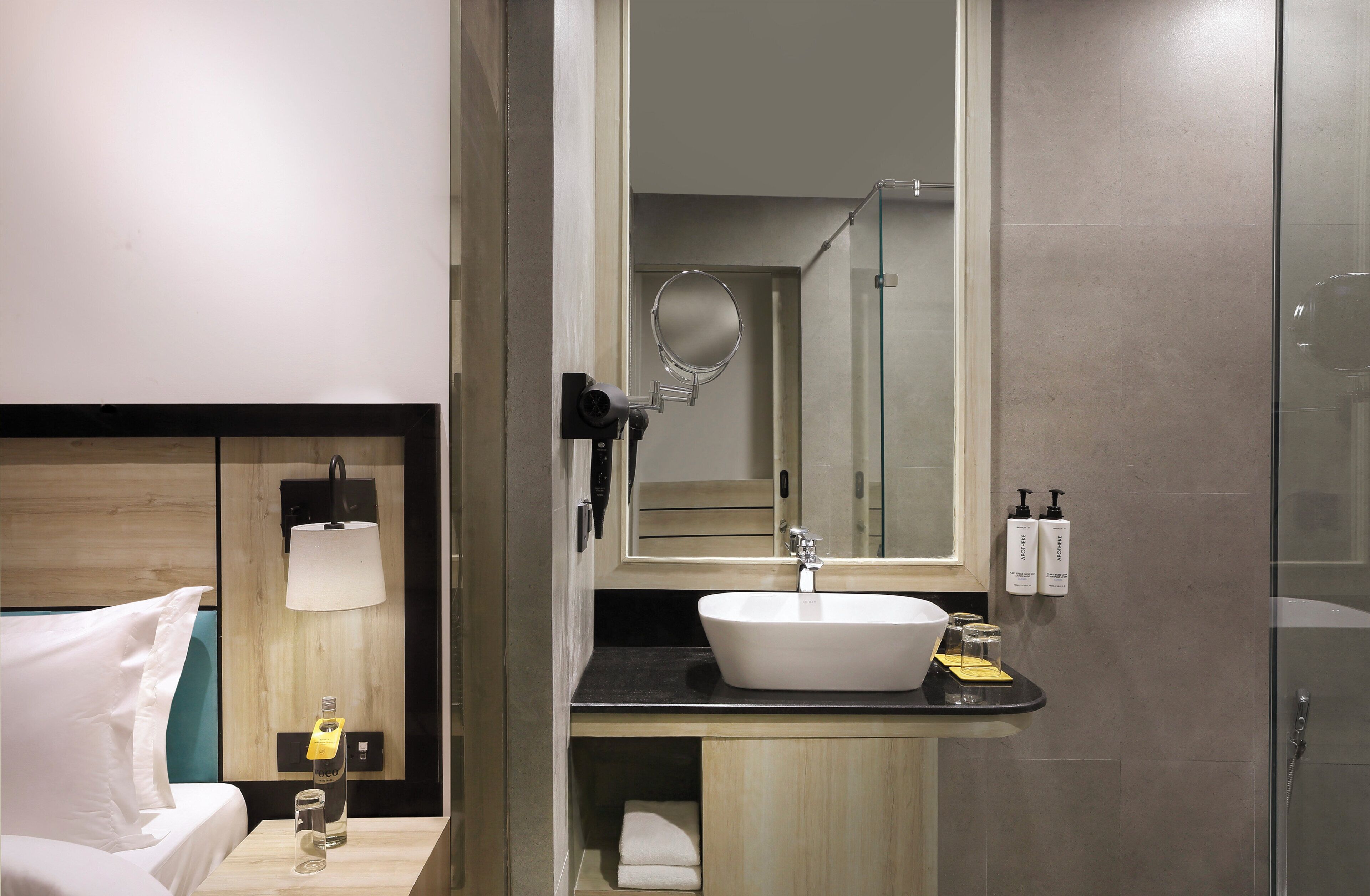 A hotel bathroom with a large mirror, a sink, and a shower. There is a towel rack and a bottle of hand soap on the counter.