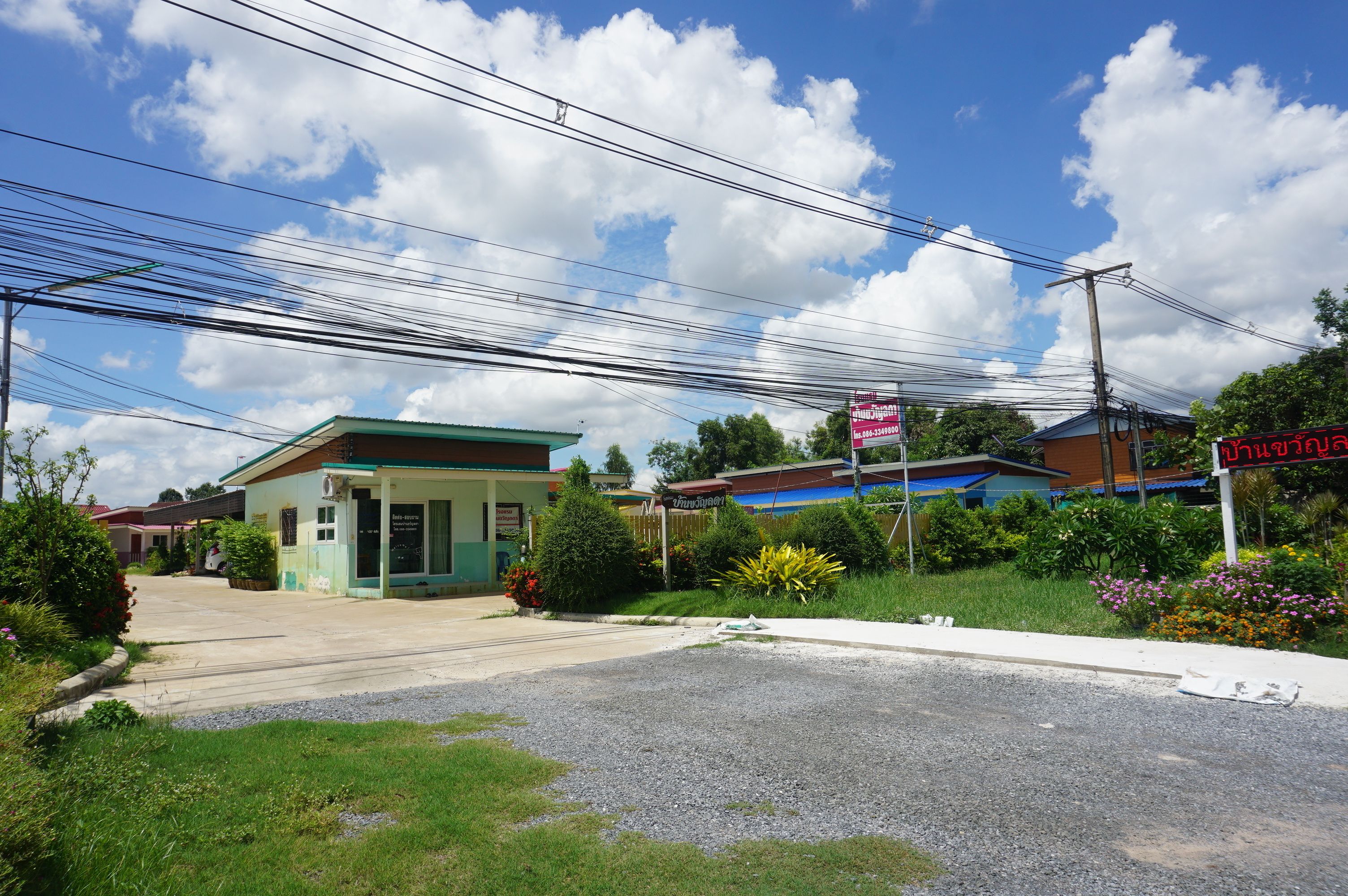 A small building with a green roof and a signboard, surrounded by greenery and a gravel road.