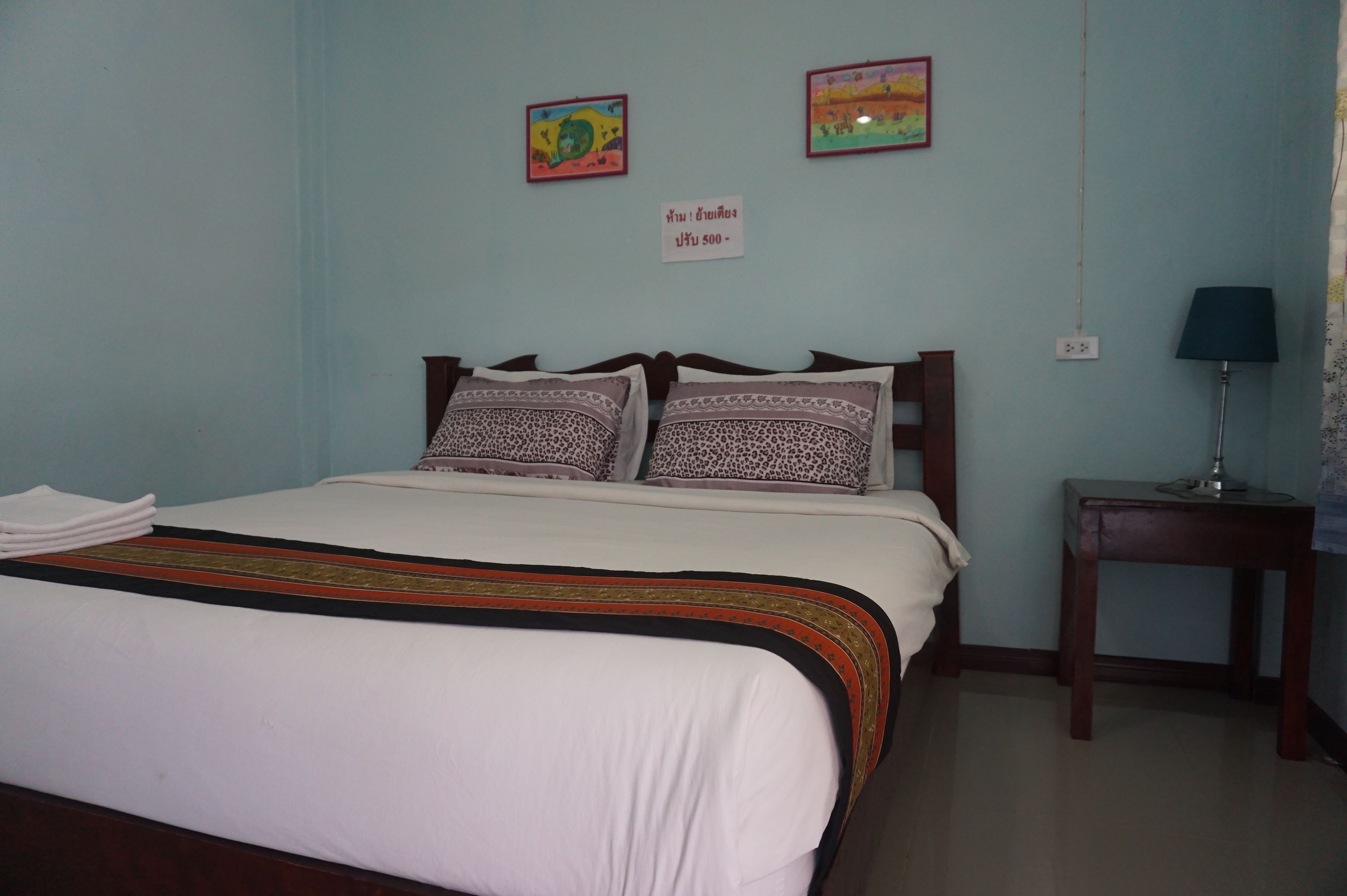 A neatly made bed with a white and patterned bedspread, two pillows, and a decorative border. There are two framed pictures on the wall, a small table with a lamp, and a sign indicating room number 500.