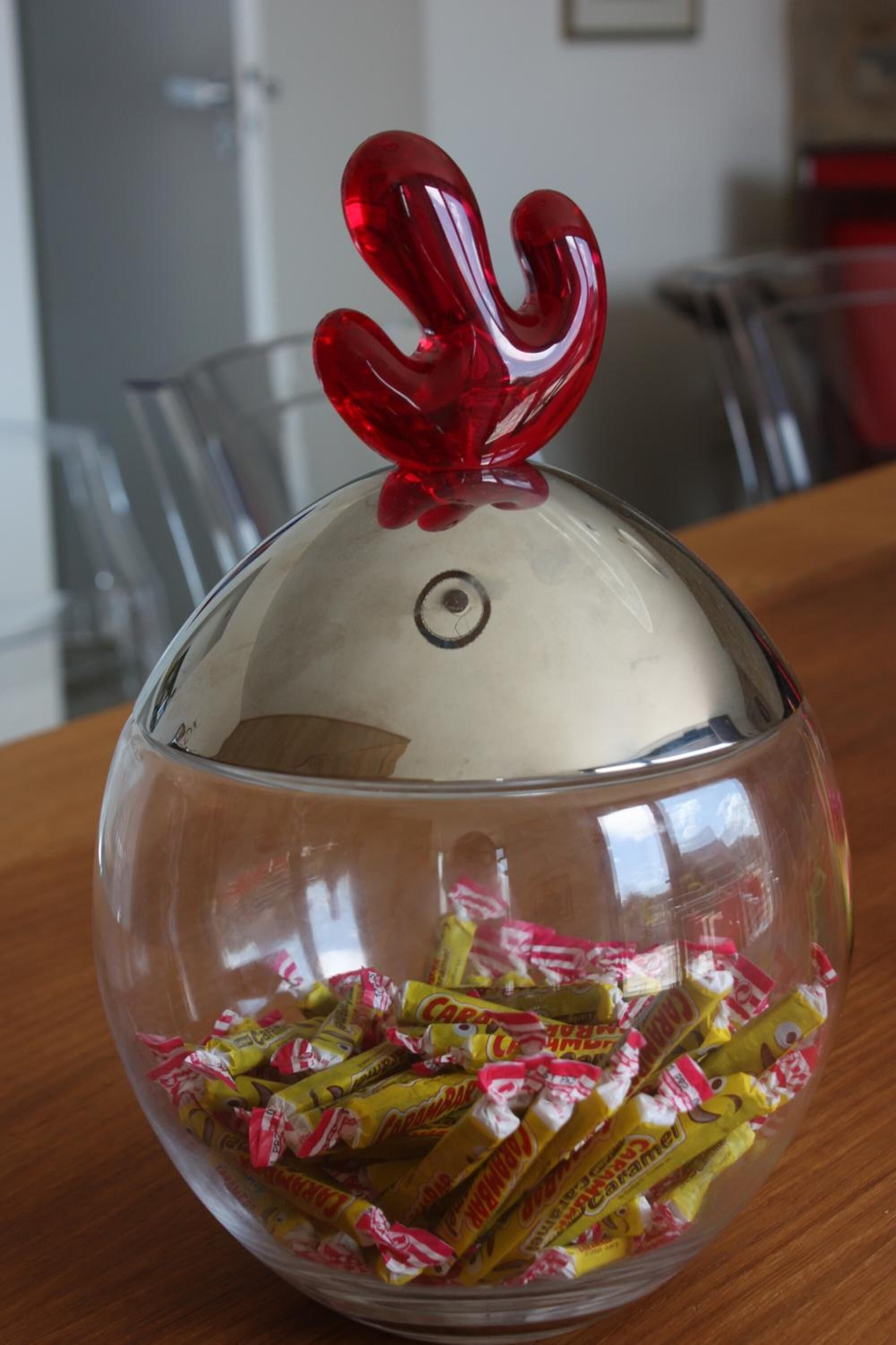 Un tazón de vidrio transparente con tapa, lleno de caramelos envueltos, sobre una superficie de madera.