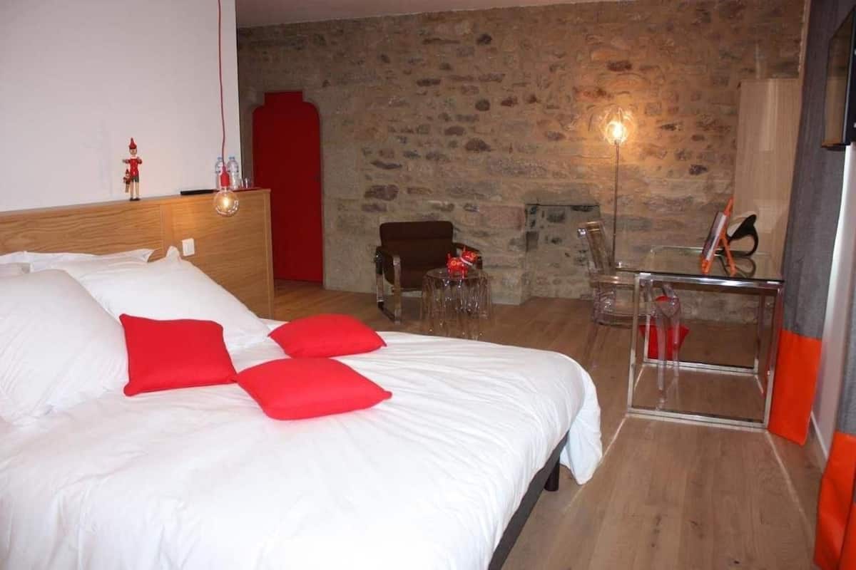 Une chambre à coucher avec une tête de lit en bois, un grand lit avec une literie blanche et rouge, un mur en pierre, une armoire rouge et une table en verre transparent agrémentée de touches orange.
