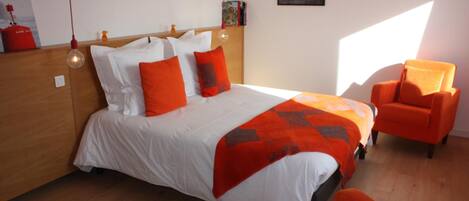 A bedroom with a wooden headboard, a bed with white and orange bedding, a chair, and a small table.