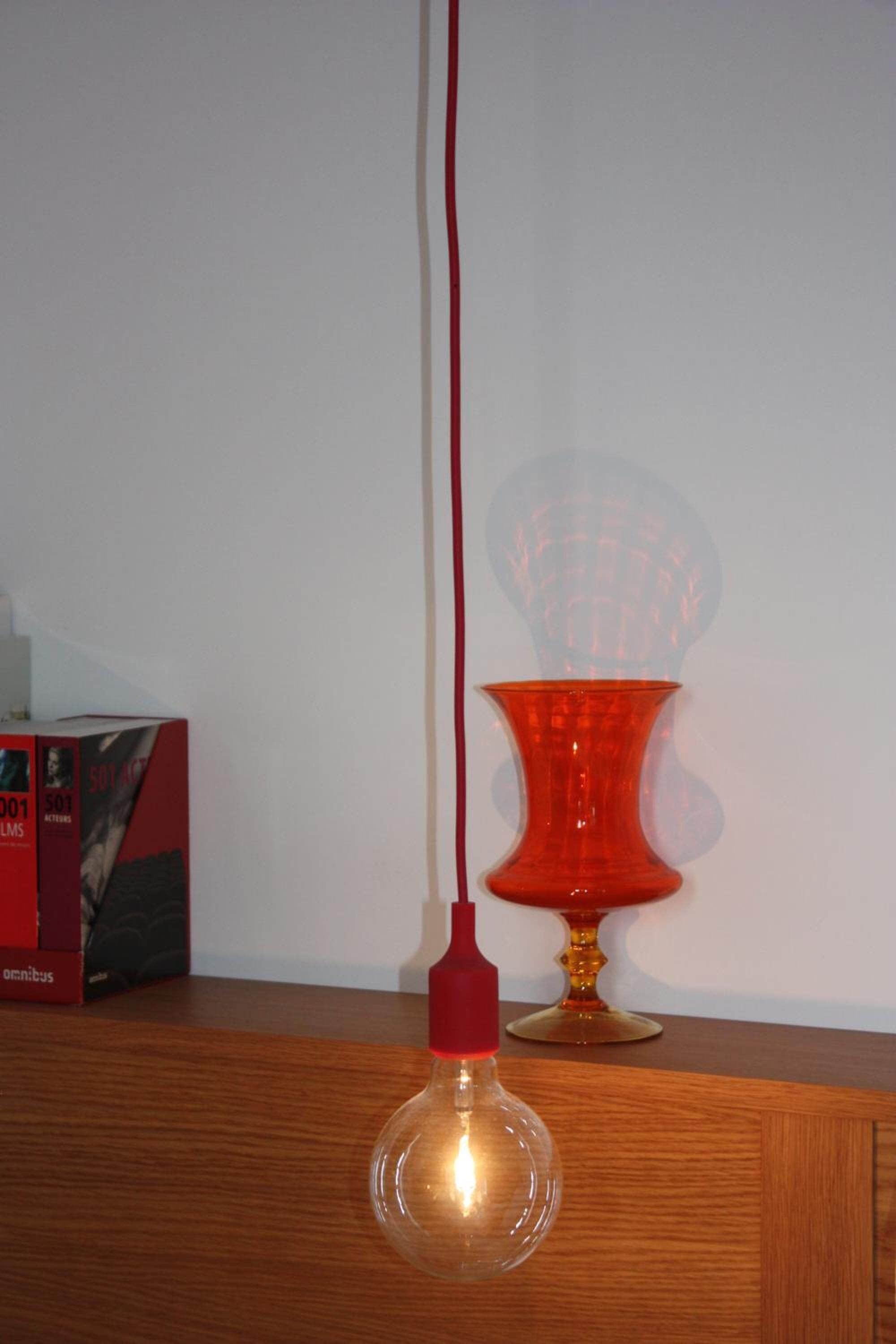 A clear glass light bulb hanging from a red cord, a red vase, and a box labeled "SOY ART" on a wooden surface.