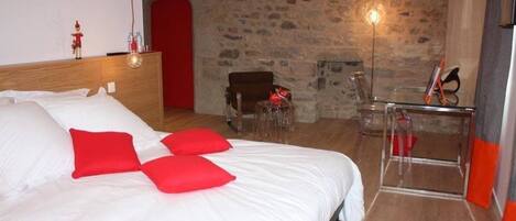 Un dormitorio con cabecera de madera, cama con ropa de cama blanca y roja, pared de piedra, una silla y una mesa de vidrio con un objeto rojo encima.