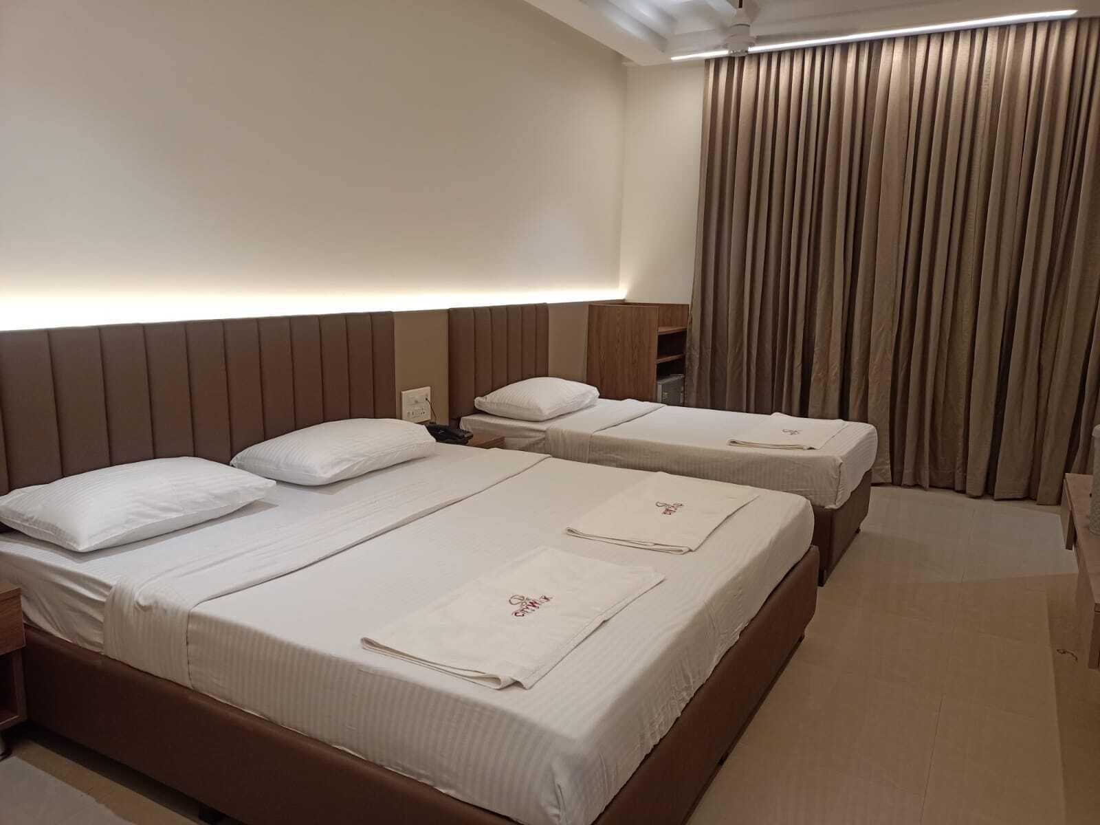 Habitación de hotel con dos camas, cada una con sábanas y almohadas blancas. Se observa un cabecero de madera, una mesita de noche y una lámpara. La habitación tiene un ventanal grande con cortinas.