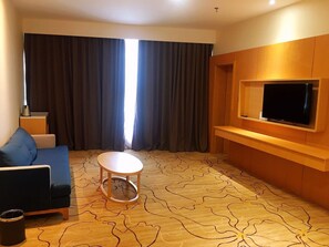 Habitación de hotel con un sofá azul, una mesita redonda pequeña, un televisor fijado a una pared de madera y un ventanal con cortinas oscuras.