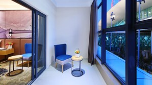 Una habitación de hotel moderna con un separador de vidrio, una silla azul, una pequeña mesa redonda con un florero y vistas a un edificio con detalles rojos.