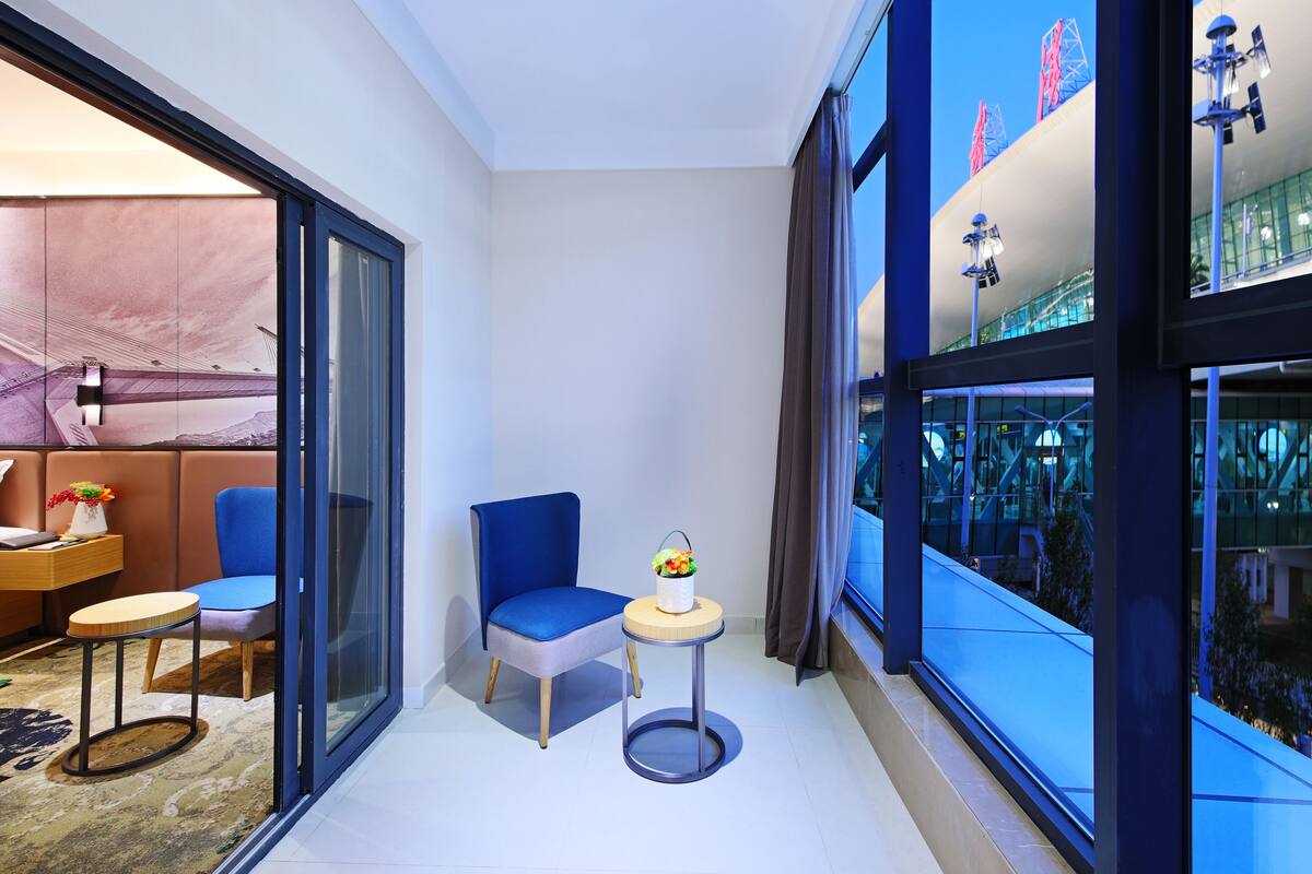 Una habitación de hotel moderna con un separador de vidrio, una silla azul, una pequeña mesa redonda con un florero y vistas a un edificio con detalles rojos.