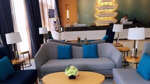 Un moderno vestíbulo de hotel con un sofá gris, cojines de acento azul, una mesa redonda de madera y un mostrador de recepción al fondo.