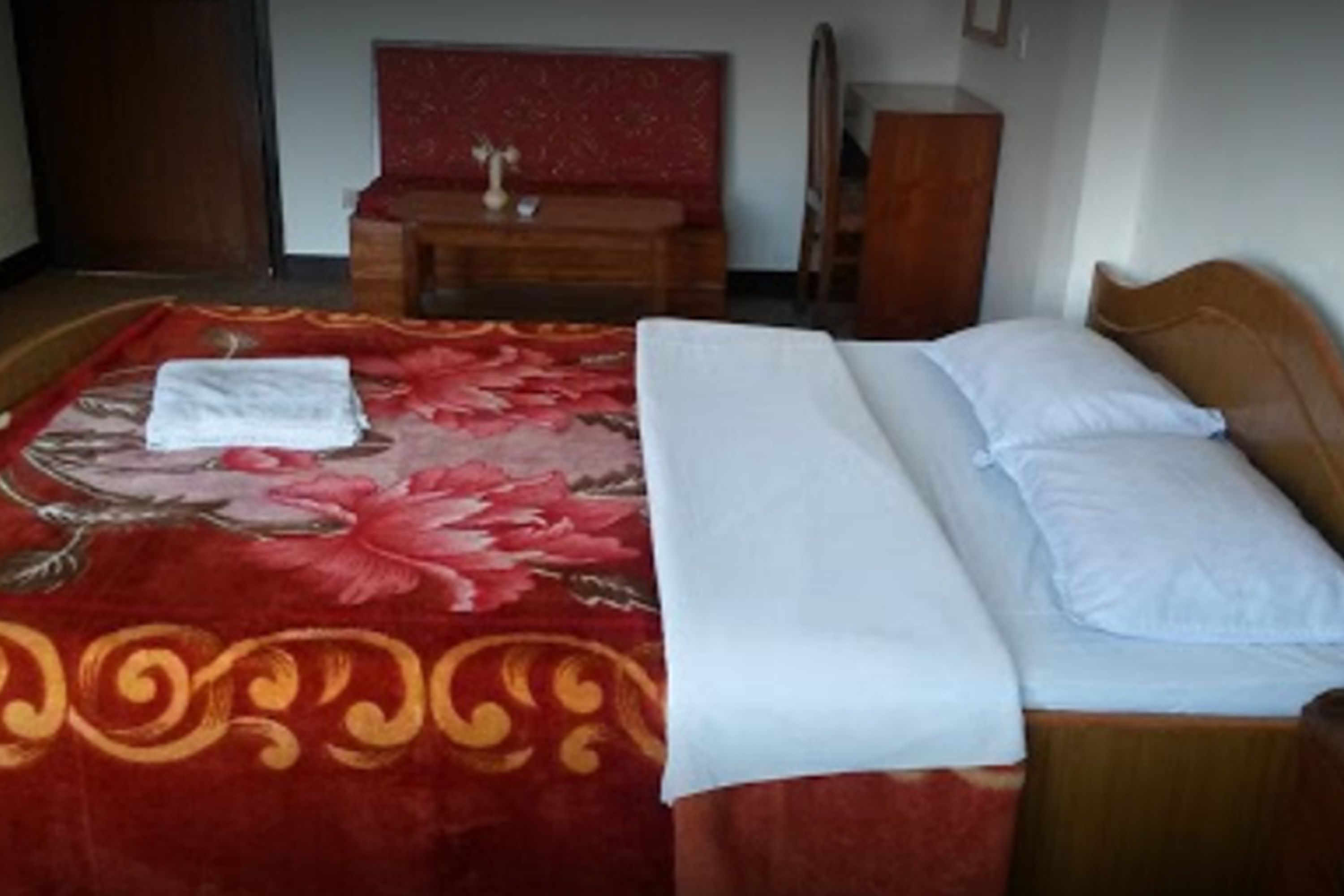A neatly made bed with a red floral bedspread and white linens, a wooden nightstand, and a small table with a vase.