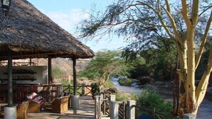 A thatched-roof outdoor seating area with wicker furniture and a view of a river and wildlife. - Man Eaters Lodge (Tsavo)