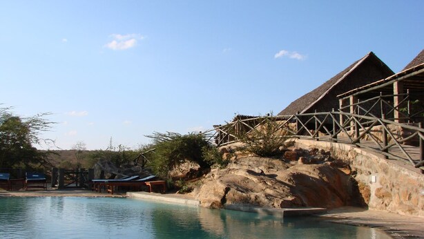 A pool area with a wooden bridge and a building with a thatched roof. - Man Eaters Lodge (Tsavo)