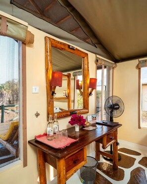 A wooden vanity with a mirror, a fan, and a small table with bottles and a vase of flowers. - Man Eaters Lodge (Tsavo)