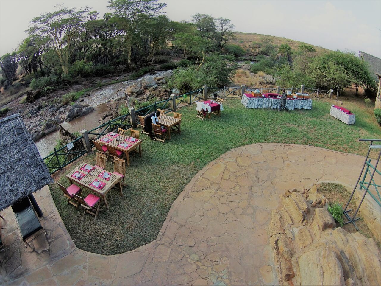 Un espace extérieur aménagé pour les repas, avec des tables et des chaises, un sentier en pierre et un paysage naturel agrémenté d’arbres et d’un ruisseau.
