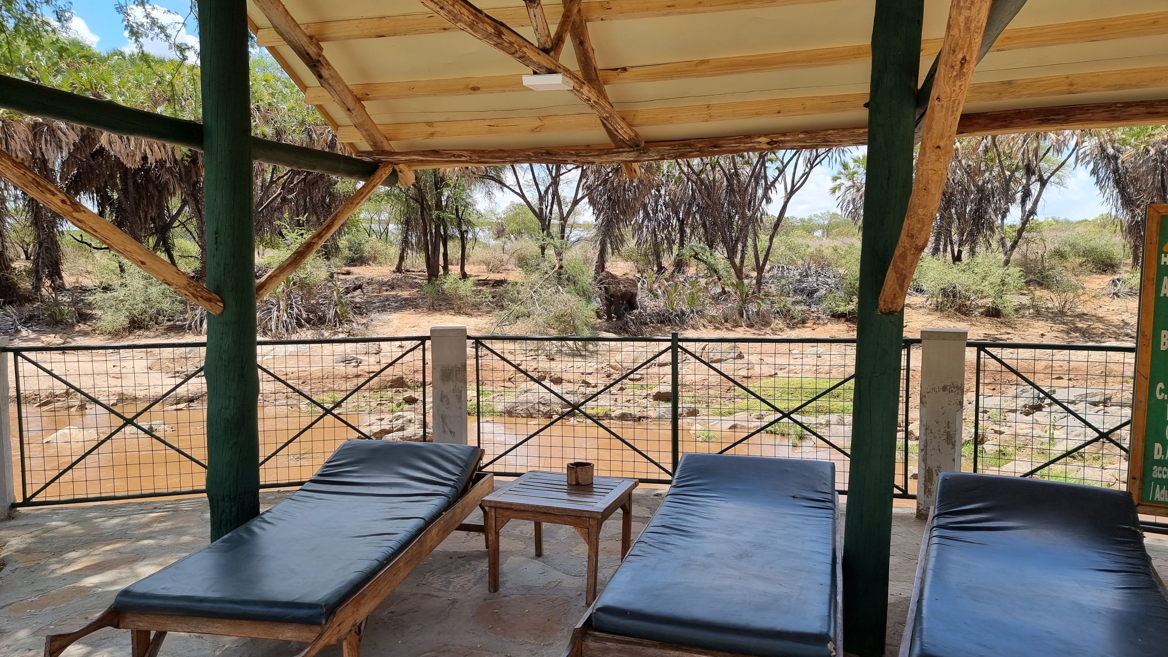 A covered outdoor seating area with two lounge chairs and a small table, overlooking a natural landscape with trees and a dirt path.