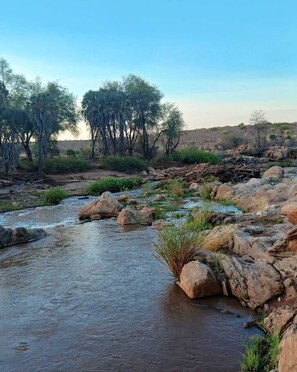 A natural landscape with a river, rocks, and sparse vegetation. - Man Eaters Lodge (Tsavo)
