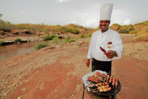 A chef grilling skewers outdoors with a river in the background. - Man Eaters Lodge (Tsavo)