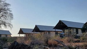 A row of cabins with sloped roofs, surrounded by dry vegetation and a large rock in the foreground. - Man Eaters Lodge (Tsavo)