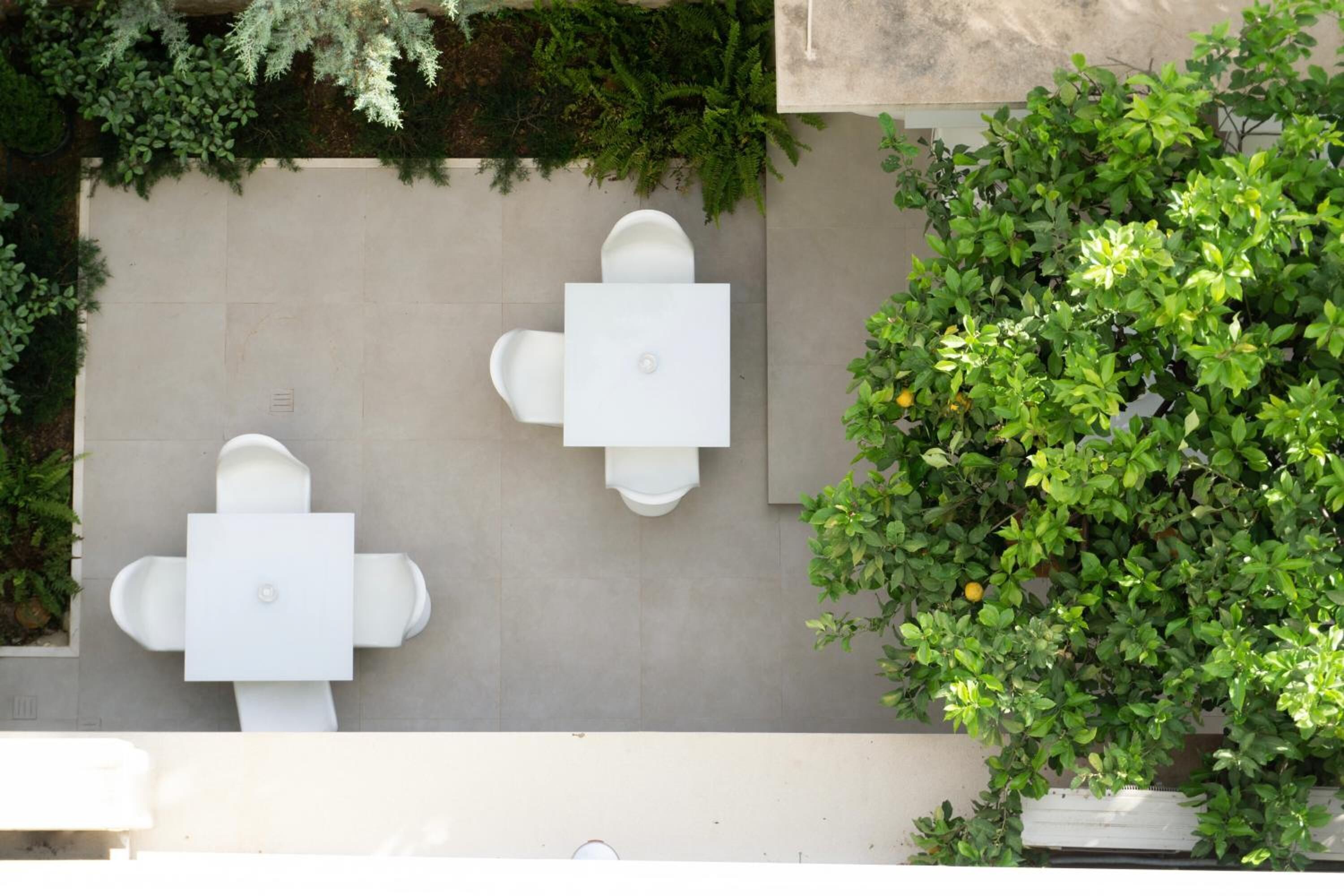 A top-down view of a patio area with two white tables and chairs, surrounded by lush greenery and a potted plant.