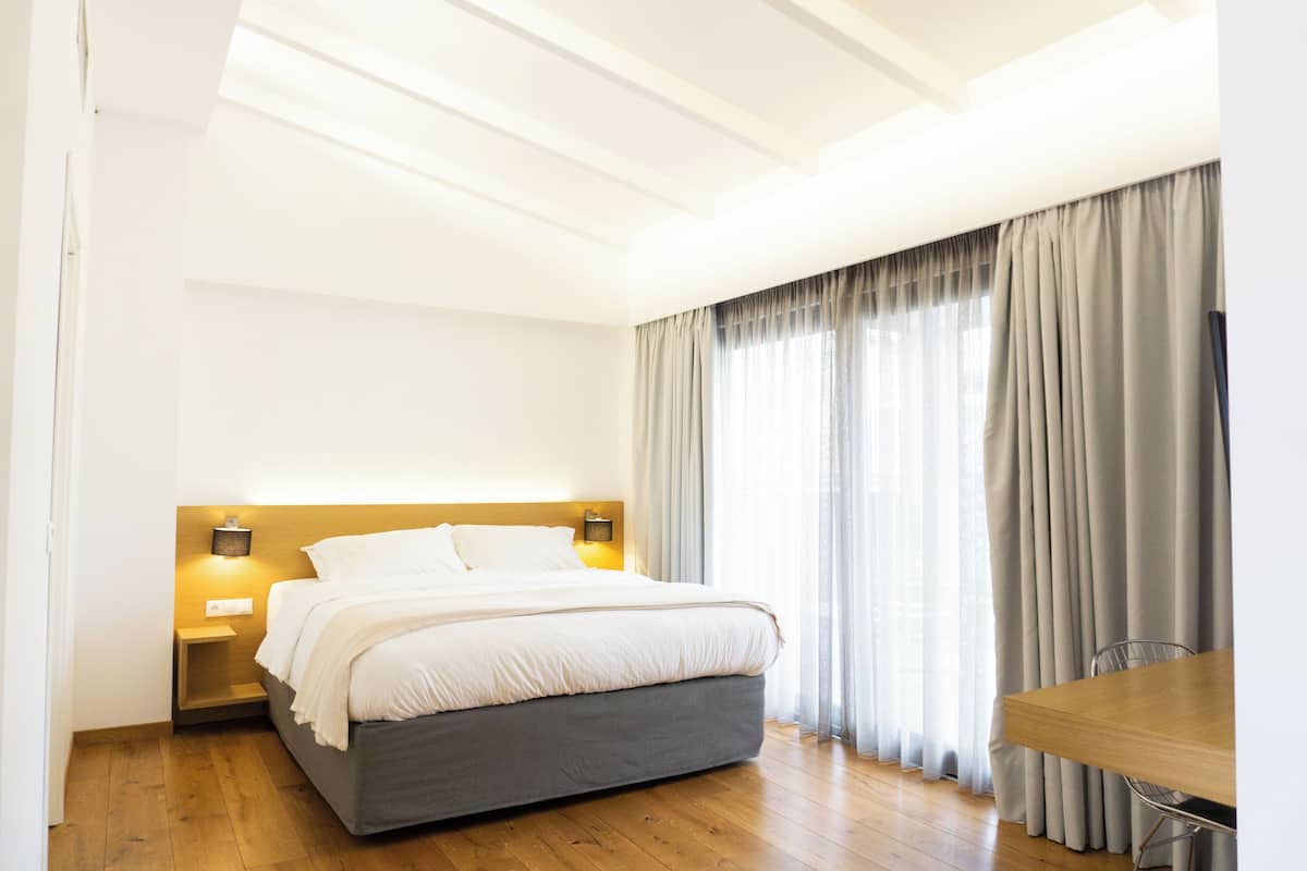 Ein modernes Schlafzimmer mit einem Bett, Nachttischlampen, einem Holztisch und einem Fenster mit Vorhängen.
