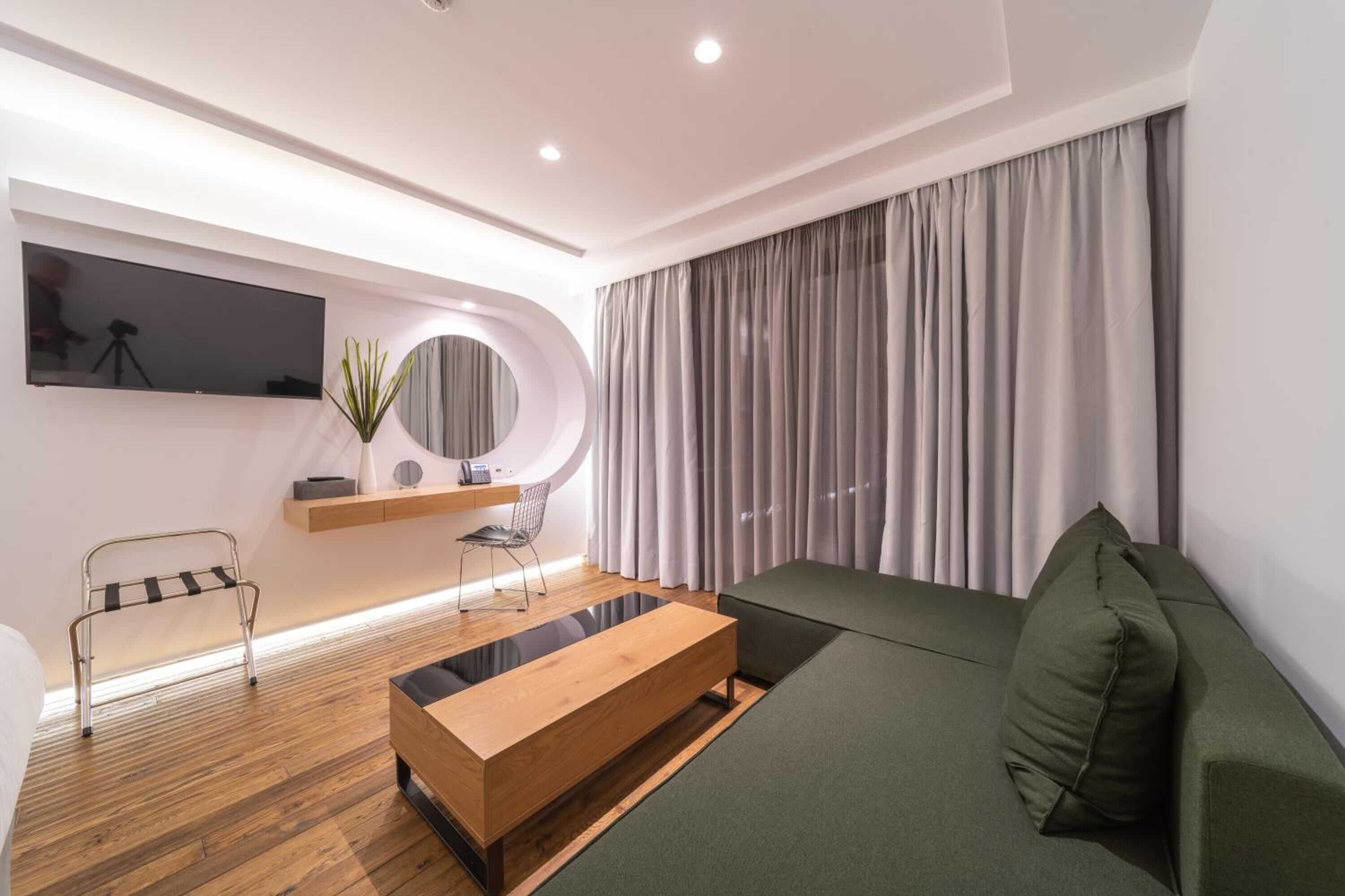 A modern living room with a wooden coffee table, a green sofa, a TV mounted on the wall, and a large window with curtains.