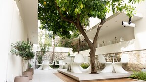 Un área de estar moderna al aire libre con sillas y mesas blancas, rodeada de plantas en macetas y un árbol.