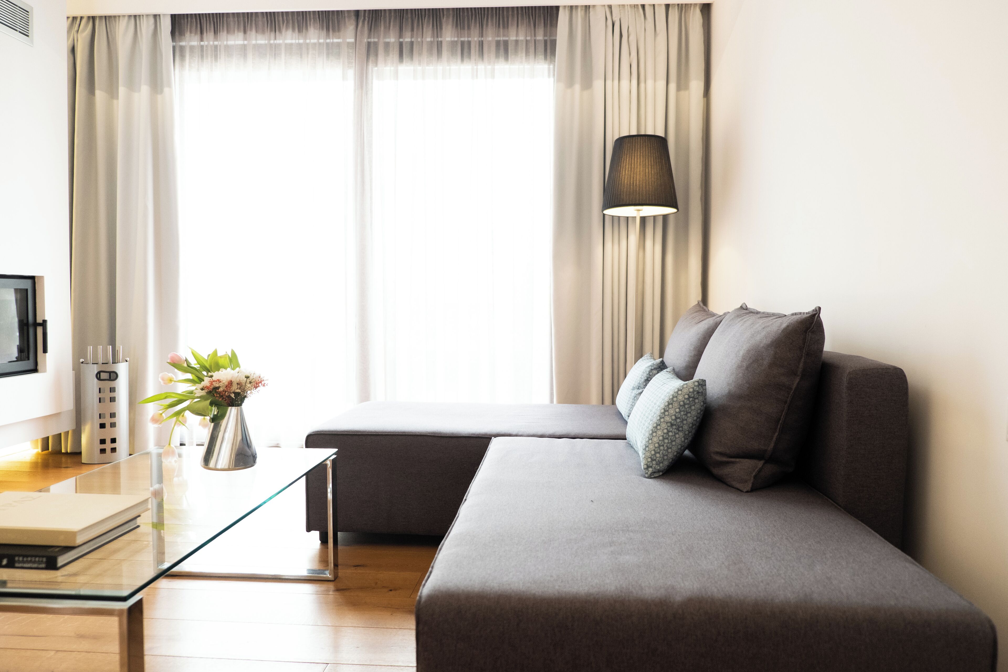 A modern living room with a grey sofa, a glass coffee table, a lamp, and a view through sheer curtains.