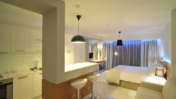 A modern hotel room with a kitchenette, a large bed, a sitting area with a sofa, and a dining area with bar stools.