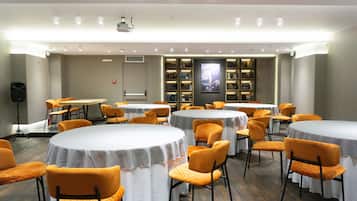 Un salón de conferencias con varias mesas y sillas, un proyector y una estantería con libros.