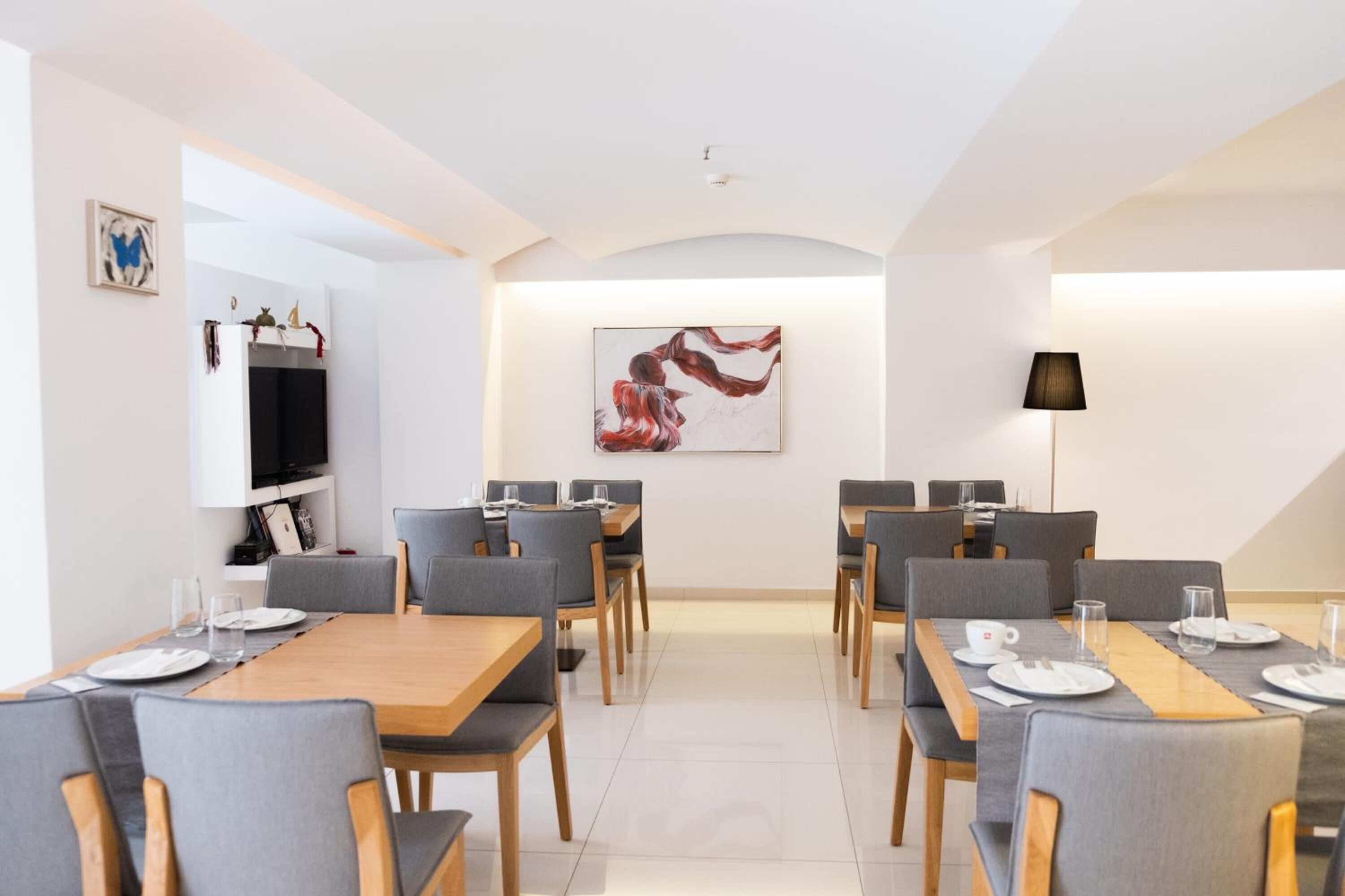 A modern dining area with wooden tables and grey upholstered chairs, a flat-screen TV mounted on the wall, and a large abstract painting above the tables.