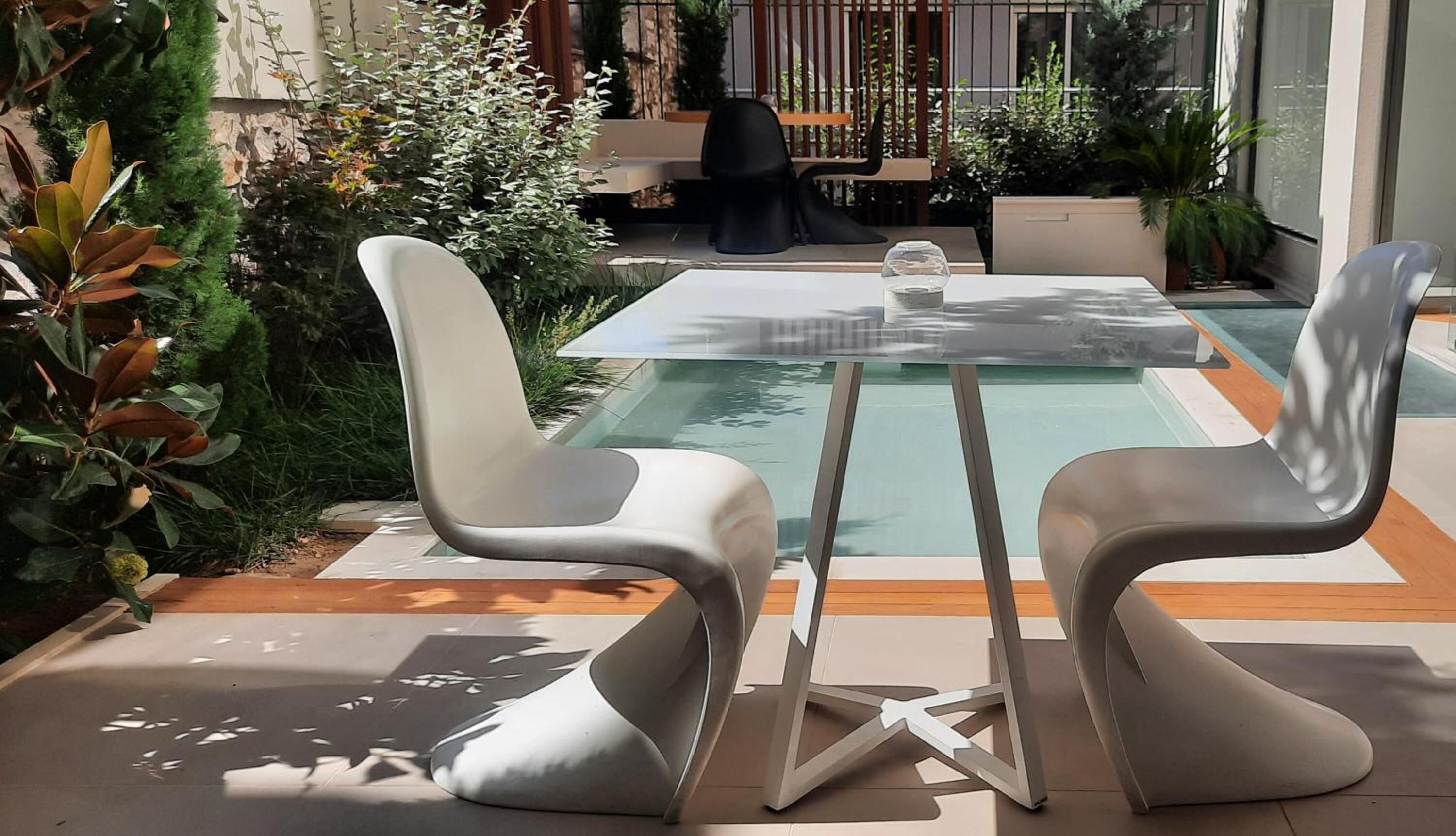 An outdoor seating area with a glass-top table and two modern chairs, surrounded by greenery and a pool.
