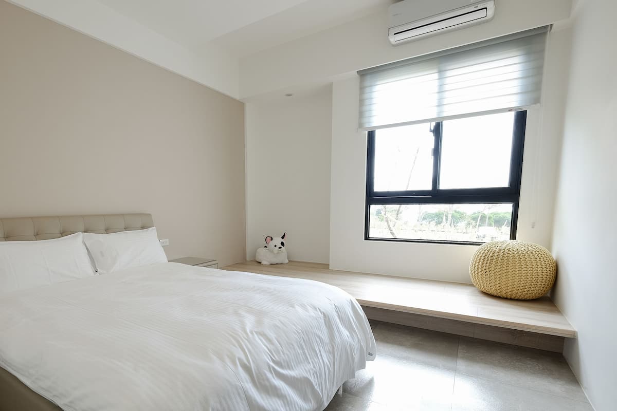 Ein Schlafzimmer mit Bett, Nachttisch, Fenster und einem kleinen Hocker.