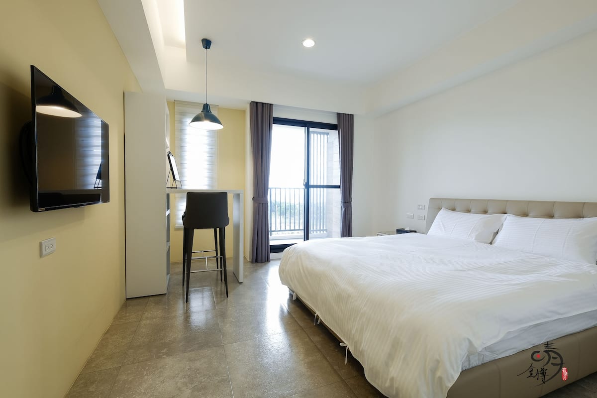 Ein Hotelzimmer mit einem großen Bett, einem Schreibtisch mit Stuhl, einem an der Wand befestigten Fernseher und einem Balkon mit Vorhängen.