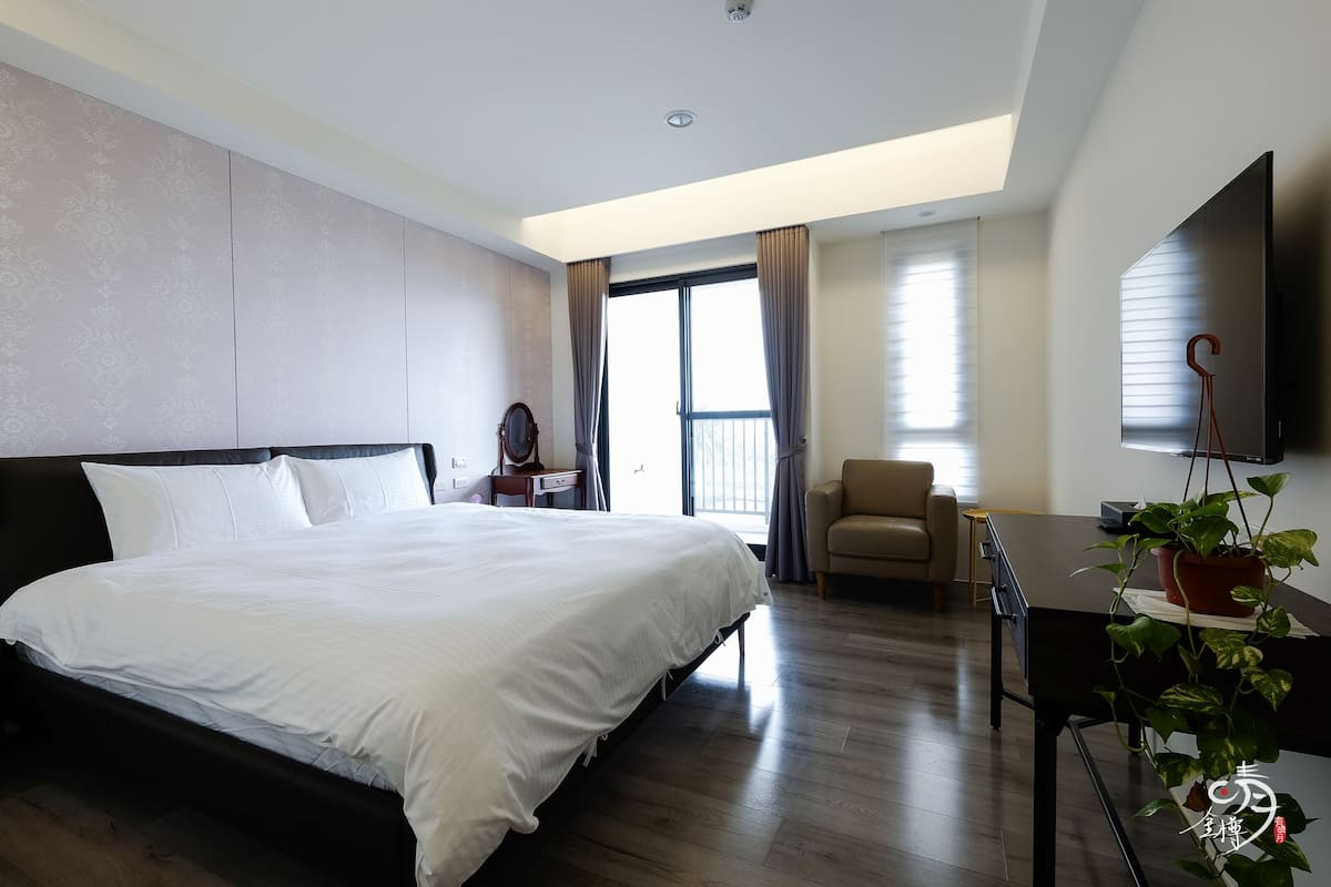 Ein modernes Hotelzimmer mit einem großen Bett, einem Schreibtisch mit Pflanze, einem Sessel und einem an der Wand befestigten Fernseher.