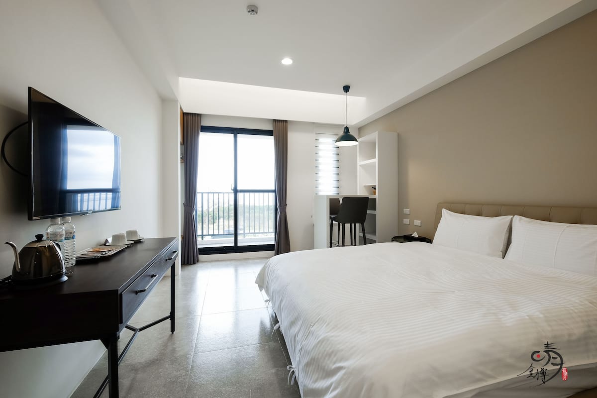 Ein Hotelzimmer mit einem großen Bett, einem Schreibtisch mit Wasserkocher, einem Flachbildfernseher und Blick ins Freie durch eine Schiebetür.