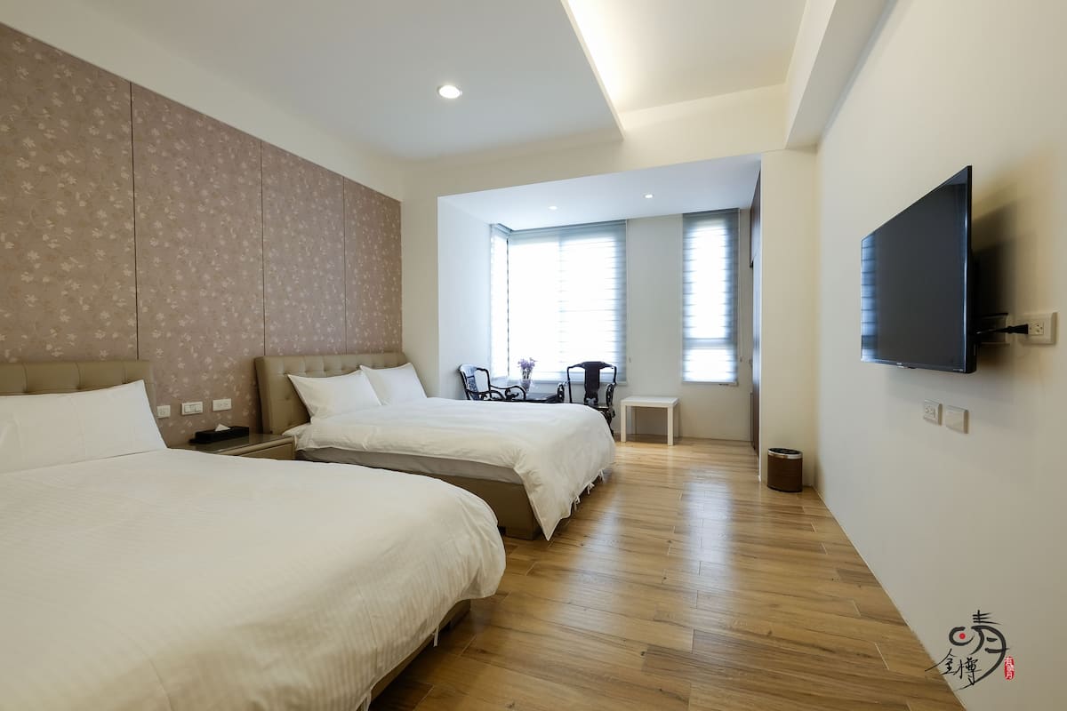 Ein Hotelzimmer mit zwei Betten, einem an der Wand montierten Fernseher und einem Fenster mit Jalousien.