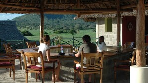 Un área de comedor al aire libre con mesas y sillas de madera, techo de paja y vistas a un paisaje verde con colinas.
