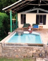 Una piscina con un muro de piedra, un porche cubierto con sillas y un edificio al fondo.