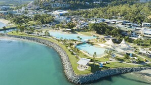 An aerial view of a coastal resort with multiple swimming pools, a rocky shoreline, and a backdrop of residential buildings. - Ocean Views 49A (Airlie Beach)