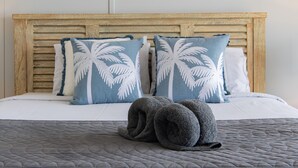 A bed with a wooden headboard, two pillows with palm leaf designs, and a rolled-up gray towel on a quilted bedspread. - Ocean Views 49A (Airlie Beach)