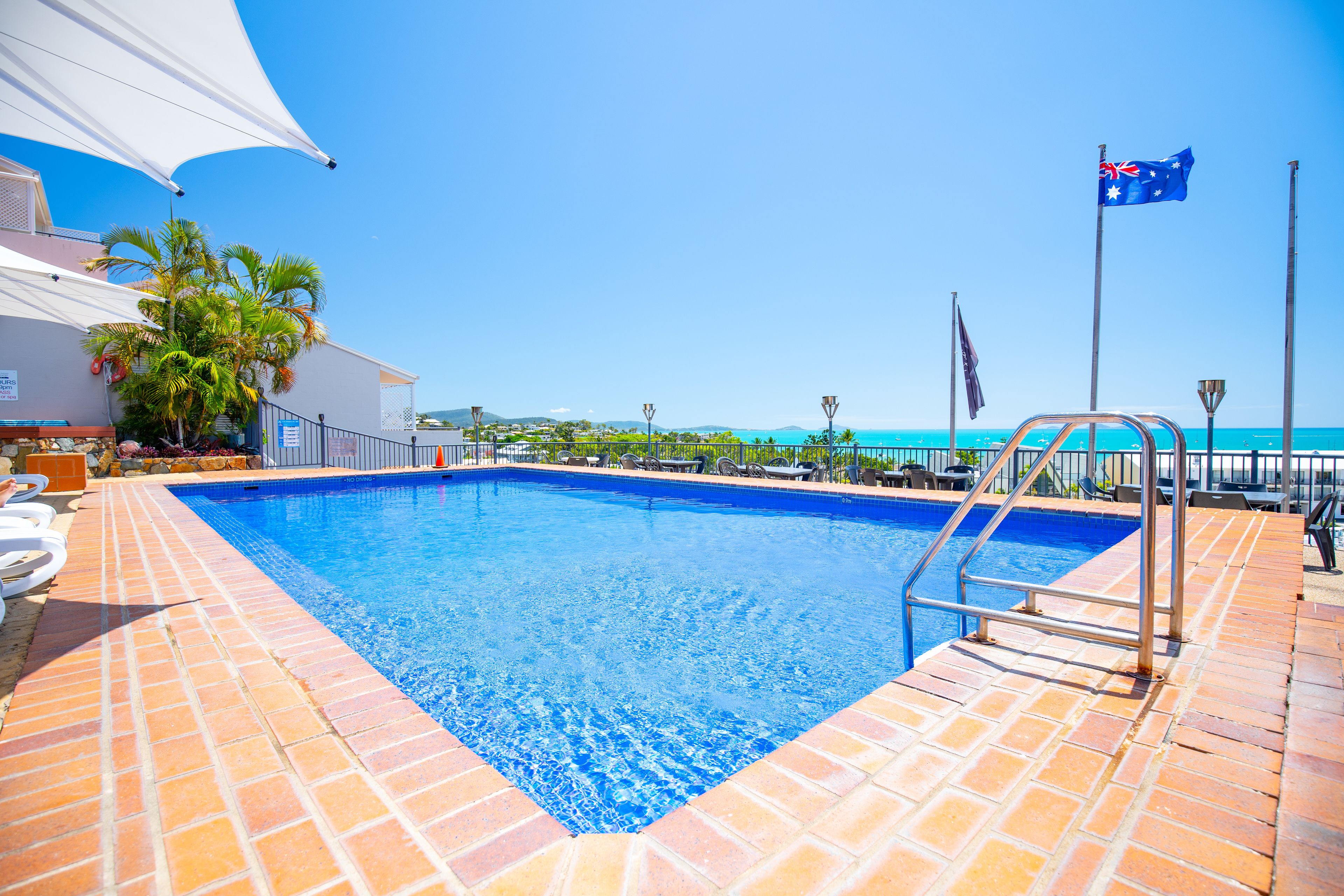 Una piscina con plataforma de baldosas, una bandera y vista al mar.