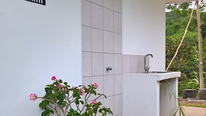 Una puerta blanca con un letrero que dice "KATITI" arriba, una planta en maceta con flores rosadas y un área de lavamanos con cabezal de ducha.