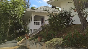 Una casa de dos pisos con una escalera que lleva a la entrada, rodeada de abundante follaje y plantas en macetas.